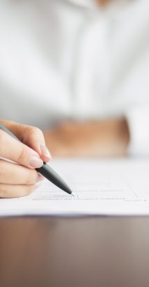 Hand of young businesswoman wearing ring and sitting at table writing on document with pen in office